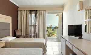 Hotellrum med två sängar, TV, bord och stolar samt en balkong med utsikt över grönska och hav. (AI-genererad text)