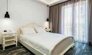 Ett sovrum med en vit säng och vita sängbord, bredvid ett stort fönster med ljusa gardiner. (AI-genererad text)