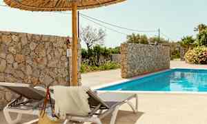 Två solstolar med en handduk och stråväska bredvid en pool. Här finns också en stenmur och palmer i bakgrunden. (AI-genererad text)