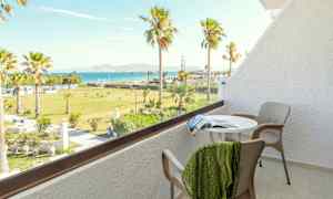 En balkong med bord, stolar och utsikt över en grön trädgård med palmer samt havet i bakgrunden. (AI-genererad text)