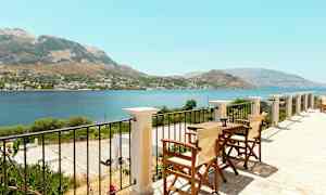 Utsikt från en terrass med bord och stolar över hav och berg. (AI-genererad text)