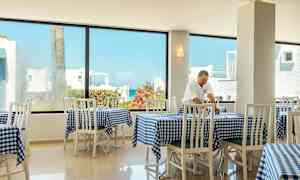 Ljus restaurang med vita stolar och rutiga blå dukar, samt stora fönster som visar utsikt över byggnader och grönska. (AI-genererad text)