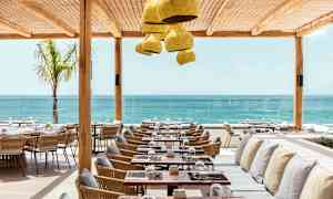 Restaurang med havsutsikt, med bord och stolar i trä och rotting samt kuddprydda bänkar under ett skyddande tak. (AI-genererad text)