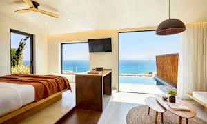 Ett rum med utsikt över havet genom stora fönster, en säng är placerad till vänster och ett bord med föremål finns i förgrunden. (AI-genererad text)