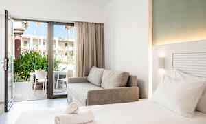 Ett modernt hotellrum med en dubbelsäng, en grå soffa och en öppen glasdörr som leder till en terrass med stolar och grönska. (AI-genererad text)