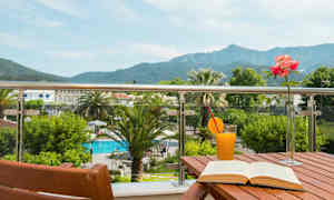 Balkong med utsikt över pool och berg, bord med bok, juice och blomvas. (AI-genererad text)