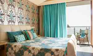 Ett rum med en säng dekorerad med blå och vita blommiga mönster, en fondvägg med blommotiv och draperier i turkos som leder till en balkong. (AI-genererad text)