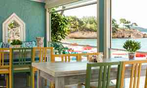 En ljus restaurang med färgade trästolar och bord, med utsikt över havet genom stora fönster. (AI-genererad text)