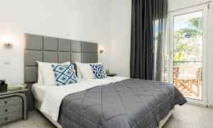 Ett sovrum med en grå sänggavel, dubbelsäng klädd med grå överkast och vita kuddar, samt färgstarka dekorativa kuddar. Rummet har ett fönster med utsikt och en liten möblerad balkong. (AI-genererad text)