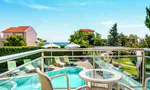 Terrass med bord och stolar som vetter mot en utomhuspool, grönska och hav i bakgrunden. (AI-genererad text)