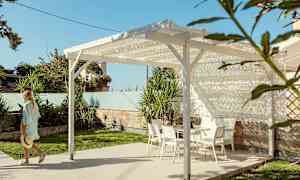 En person går intill en pergola med vita trädgårdsmöbler på en stenlagd yta, omgiven av grönska. (AI-genererad text)