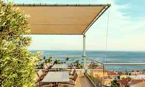 Terrass med bord och stolar, ett tak av tyg och utsikt över havet och hus. (AI-genererad text)