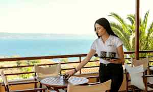 En person placerar föremål på ett bord på en utomhusterrass med havsutsikt. (AI-genererad text)