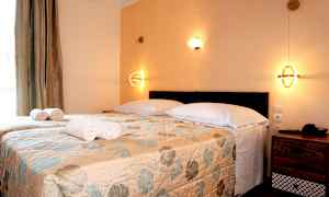 Ett sovrum med två enkelsängar täckta med ett blommigt överkast, vita kuddar och vikta handdukar, samt nattlampor på väggen. (AI-genererad text)