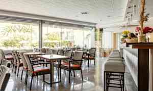 En hotellrestaurang med bord och stolar samt en bardisk, stora fönster mot utomhus. (AI-genererad text)