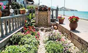 En liten fontän omgiven av blommor och växter vid havet. (AI-genererad text)