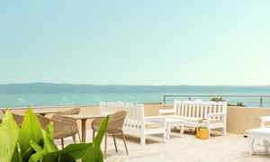 Utemöbler med bord och stolar i ljusa färger på en terrass med havsutsikt. (AI-genererad text)