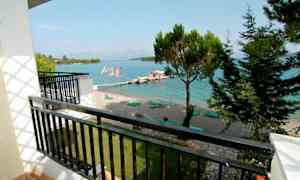 Utsikt från en balkong mot en strand med solstolar vid en klarblå havsvik. Träd och moln i bakgrunden. (AI-genererad text)