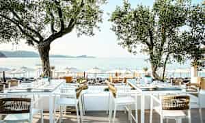 Uteservering med bord och stolar under träd, utsikt mot havet och parasoller på stranden. (AI-genererad text)