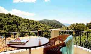 Utsikt från en balkong med bord, dryck och en stol med en duk, samt träd och berg i bakgrunden. (AI-genererad text)