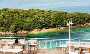 Två personer sitter vid bord på en terrass med utsikt över havet och en grönskande kustlinje. (AI-genererad text)