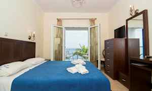 Hotellrum med dubbelsäng, blå överkast, och utsikt mot havet genom öppna glasdörrar. (AI-genererad text)