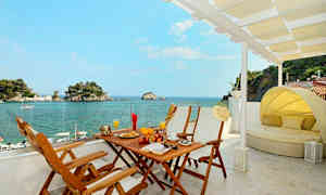 Utsikt från en terrass med matbord och solsäng mot havet och små öar. (AI-genererad text)