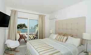 Ett hotellrum med en stor säng, beige inredning och utsikt över havet från en balkong. (AI-genererad text)