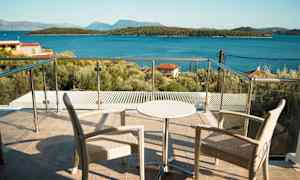 Två stolar och ett mindre bord på en terrass med glasräcke, utsikt mot havet och skogklädda öar. (AI-genererad text)