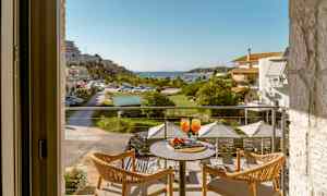 Utsikt från en balkong med två stolar och ett bord som har två glas med orange dryck, mot en grönskande terrass och en hamn. (AI-genererad text)