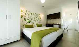 Ett hotellrum med en dubbelsäng täckt med vit sängkläder och gröna filtar, en garderob, ett pentry och en dörr i bakgrunden. (AI-genererad text)