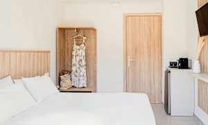 Ett hotellrum med en vit säng, en garderob med en klänning och väska, en dörr och ett litet köksområde med en kaffemaskin. (AI-genererad text)