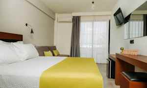 Ett hotellrum med en dubbelsäng med vita sängkläder och gult överkast, en soffa med kuddar, ett skrivbord med spegel, och en TV på väggen. Balkongdörrar med ljusa gardiner. (AI-genererad text)