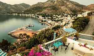 Utsikt från terrassen över en hamn med båtar, omgivande byggnader och berg i bakgrunden. (AI-genererad text)