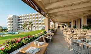 En utomhusrestaurang med dukade bord, grönska och en flerfärgad byggnad i bakgrunden. (AI-genererad text)