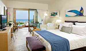 Ett hotellrum med dubbelsäng, soffa och stor fönsterdörr som leder till en terrass med utsikt över havet. (AI-genererad text)