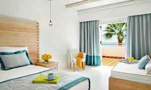 Ett ljust och luftigt hotellrum med säng, träpaneler, blommor på ett sidobord och öppna glasdörrar som leder till en balkong med havsutsikt. (AI-genererad text)