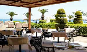 Uteservering med dukade bord, palmer och utsikt mot havet. (AI-genererad text)