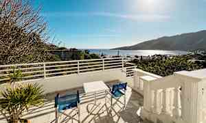 Utsikt från en terrass med bord och stolar, med hav och berg i bakgrunden. (AI-genererad text)