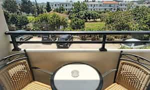 En balkong med två stolar och ett runt bord med en askkopp, med utsikt över en parkering och grönska. (AI-genererad text)