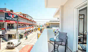 En balkong med bord, stol och blommor i förgrunden, samt en vy över en gata med butiker och byggnader. (AI-genererad text)