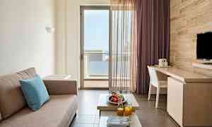 Ett rum med en beige soffa, ett bord med frukt och juice samt en balkongdörr med utsikt. (AI-genererad text)