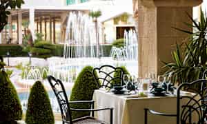 En dukad matplats utomhus med utsikt över en trädgård med fontäner. (AI-genererad text)