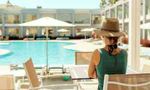 En person sitter vid ett bord nära en pool med parasoller. I bakgrunden syns en hotellbyggnad. (AI-genererad text)