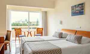 Ett hotellrum med två enkelsängar, en sittgrupp och en utsikt mot en grönskande omgivning. (AI-genererad text)