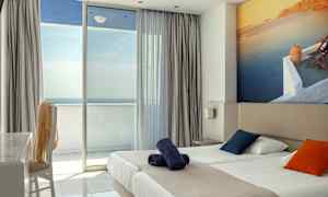 Hotellrum med en dubbelsäng, färgglada kuddar och en balkong med havsutsikt. (AI-genererad text)