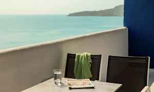 Två stolar med en grön filt på den ena, stående vid ett bord som har ett glas vatten och en tidning, med havsutsikt. (AI-genererad text)