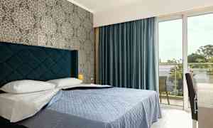 Ett hotellrum med en säng, blått överkast och blå gardiner, samt ett fönster som leder till en balkong. (AI-genererad text)