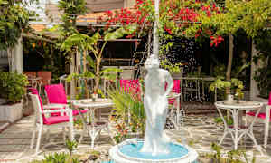 En fontän med en vit staty av en kvinna i en lummig trädgård med bord och stolar. (AI-genererad text)