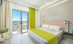 Ett hotellrum med en säng, grönt överkast, ljusa möbler och en balkong med havsutsikt. (AI-genererad text)